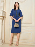 A woman wears the Social Queen Elegant Hand-Beaded Green Crystal Dress, featuring a blue, knee-length silhouette with short sleeves and green crystal embellishments, styled with a small woven handbag and strappy heels against a beige wall.