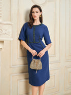 A woman wearing the Social Queen Elegant Hand-Beaded Green Crystal Dress stands indoors, holding a small textured beige handbag against an ornate cream-colored wall.