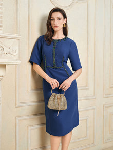 A woman wearing the Social Queen Elegant Hand-Beaded Green Crystal Dress stands indoors, holding a small textured beige handbag against an ornate cream-colored wall.