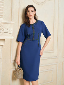 A woman wearing the Social Queen Elegant Hand-Beaded Green Crystal Dress stands by a cream ornate wall, holding a beige handbag in her left hand.