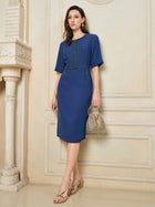 A woman stands indoors wearing the Social Queen Elegant Hand-Beaded Green Crystal Dress, featuring green crystal embellishments and a fitted waist, paired with gold strappy heels and a small woven handbag.