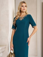 A woman in the Elegant Rhinestone-Embellished Dress by Social Queen, featuring a fitted teal chiffon silhouette with gold lace accents on the neckline and sleeves, holds a gold handbag while standing indoors against a neutral wall.