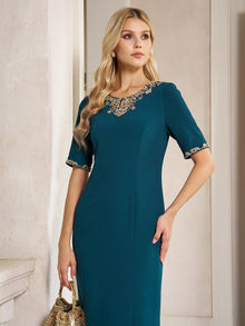 A woman in the Elegant Rhinestone-Embellished Dress by Social Queen, featuring a fitted teal chiffon silhouette with gold lace accents on the neckline and sleeves, holds a gold handbag while standing indoors against a neutral wall.