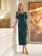 A woman in the Social Queen Elegant Rhinestone-Embellished Dress stands indoors by a staircase and classical painting, holding a woven handbag. Sparkling rhinestones accentuate the sophisticated, fitted sheath design.