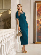 A woman wears the Social Queen Elegant Rhinestone-Embellished Dress, featuring teal chiffon and a beaded neckline, leaning against a wall with a small woven handbag in an elegant interior with classical art.
