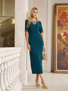 A woman stands indoors by a white balustrade and large framed painting, wearing the Social Queen Elegant Rhinestone-Embellished Dress in teal with a fitted knee-length silhouette and sparkling neckline, paired with a small handbag.