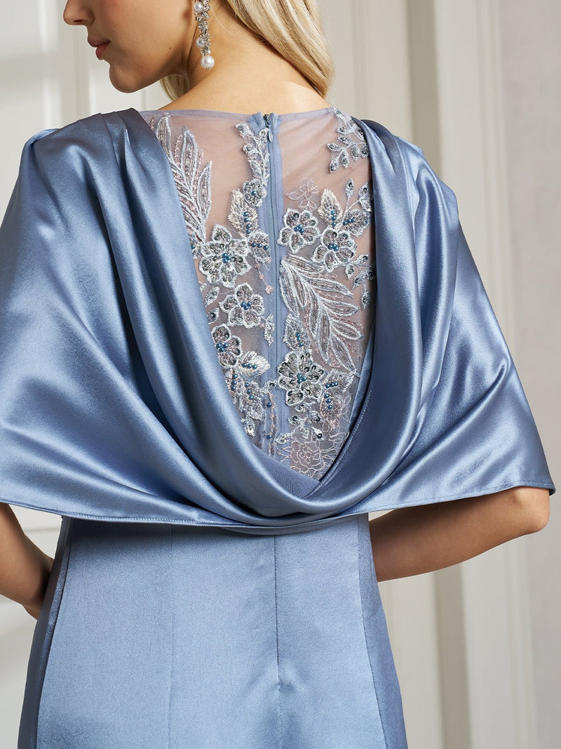 A woman wears the Social Queen Embroidered Cape-Sleeve Satin Dress, featuring light blue satin with floral embroidery and a draped back—perfect for weddings or formal events—while standing indoors.