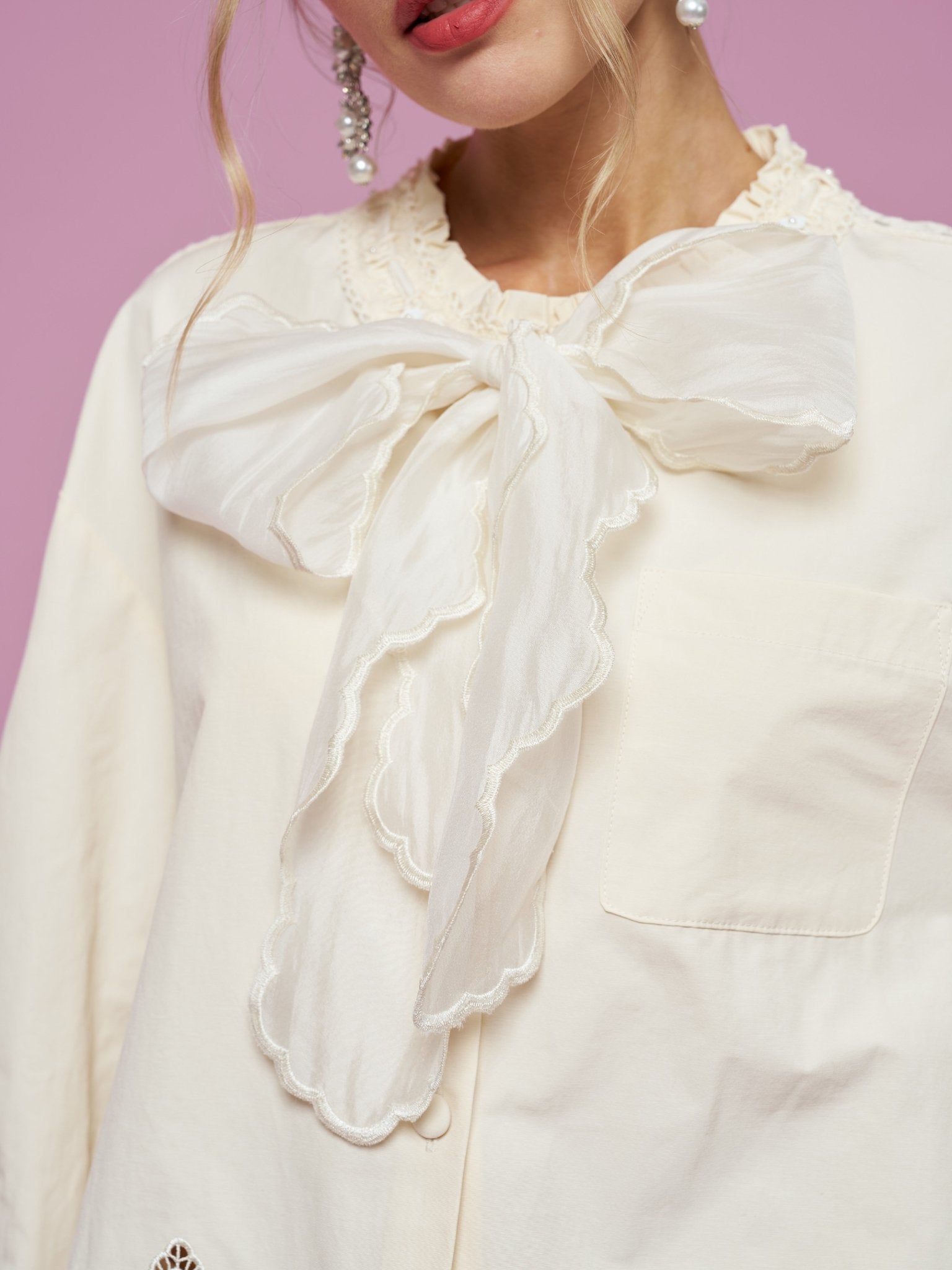 A woman, her head partially out of frame, stands against a pink background wearing the Social Queen Embroidered Cutout Bow-Tie Blouse, featuring a large sheer bow tie neckline and delicate lace accents.