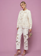 A woman stands against a pink background wearing the Social Queen Embroidered Cutout Bow-Tie Blouse, matching cutout embroidery pants, metallic sandals, and holding a small purse.
