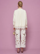A person stands facing away, wearing the Social Queen Embroidered Cutout Bow-Tie Blouse and embellished floral pants, holding a small bag, set against a pink background.