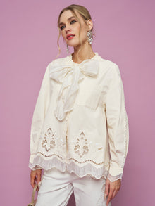 A woman stands against a pink background wearing the Embroidered Cutout Bow-Tie Blouse by Social Queen in cream, paired with white pants.