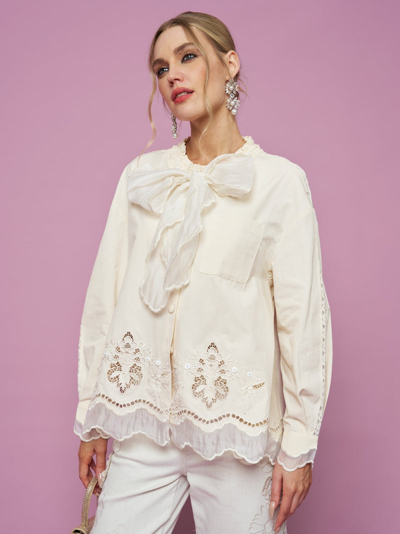 A woman stands against a pink background wearing the Embroidered Cutout Bow-Tie Blouse by Social Queen in cream, paired with white pants.