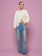 A woman stands against a pink background wearing the Social Queen Embroidered Cutout Bow-Tie Blouse, light blue wide-leg jeans with floral cutout embroidery, silver heels, drop earrings, and holding a small purse.