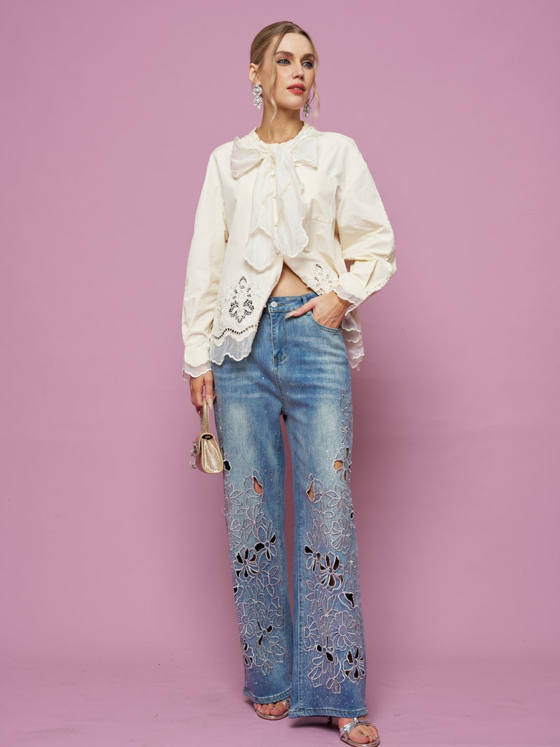 A woman stands against a pink background wearing the Social Queen Embroidered Cutout Bow-Tie Blouse, light blue wide-leg jeans with floral cutout embroidery, silver heels, drop earrings, and holding a small purse.