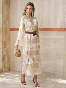 A woman in Social Queen’s Embroidered Fringe Lace Crystal-Trimmed Dress, paired with a brown belt, woven bag, and high-heeled sandals, stands by a beige stone wall—an elegant choice for a wedding guest.