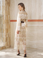 A woman stands indoors wearing the Embroidered Fringe Lace Crystal-Trimmed Dress by Social Queen, styled with a black belt, statement earrings, and high-heeled sandals—a chic choice for a stylish wedding guest.