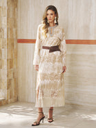 A woman stands indoors in the Social Queen Embroidered Fringe Lace Crystal-Trimmed Dress with long sleeves, a brown belt, and black heeled sandals—an elegant, neutral look ideal for wedding guests.