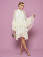 A woman stands against a pink background holding a small handbag, wearing the Social Queen Embroidered High-Neck Silk Dress featuring flared sleeves, knee-length ruffles, and subtle sequin embroidery.