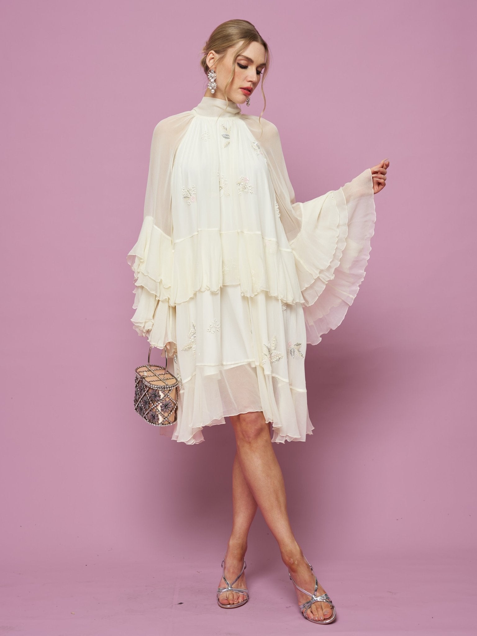 A woman stands against a pink background holding a small handbag, wearing the Social Queen Embroidered High-Neck Silk Dress featuring flared sleeves, knee-length ruffles, and subtle sequin embroidery.