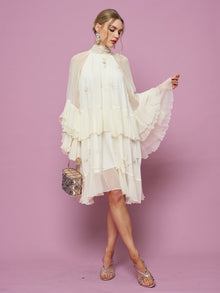 A woman stands against a pink background holding a small handbag, wearing the Social Queen Embroidered High-Neck Silk Dress featuring flared sleeves, knee-length ruffles, and subtle sequin embroidery.