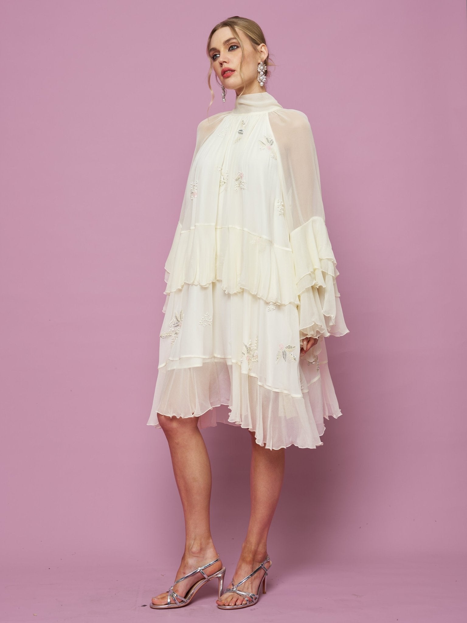 A woman poses against a pink background in Social Queen's Embroidered High-Neck Silk Dress, featuring layered off-white mulberry silk, sheer sleeves, subtle sequin embroidery, and metallic strappy heels.