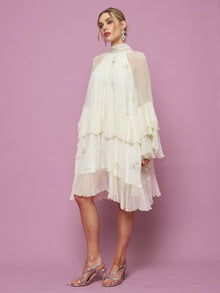 A woman poses against a pink background in Social Queen's Embroidered High-Neck Silk Dress, featuring layered off-white mulberry silk, sheer sleeves, subtle sequin embroidery, and metallic strappy heels.