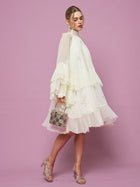 A woman wears the Social Queen Embroidered High-Neck Silk Dress with sheer sleeves and holds a small patterned handbag, standing against a pink background.