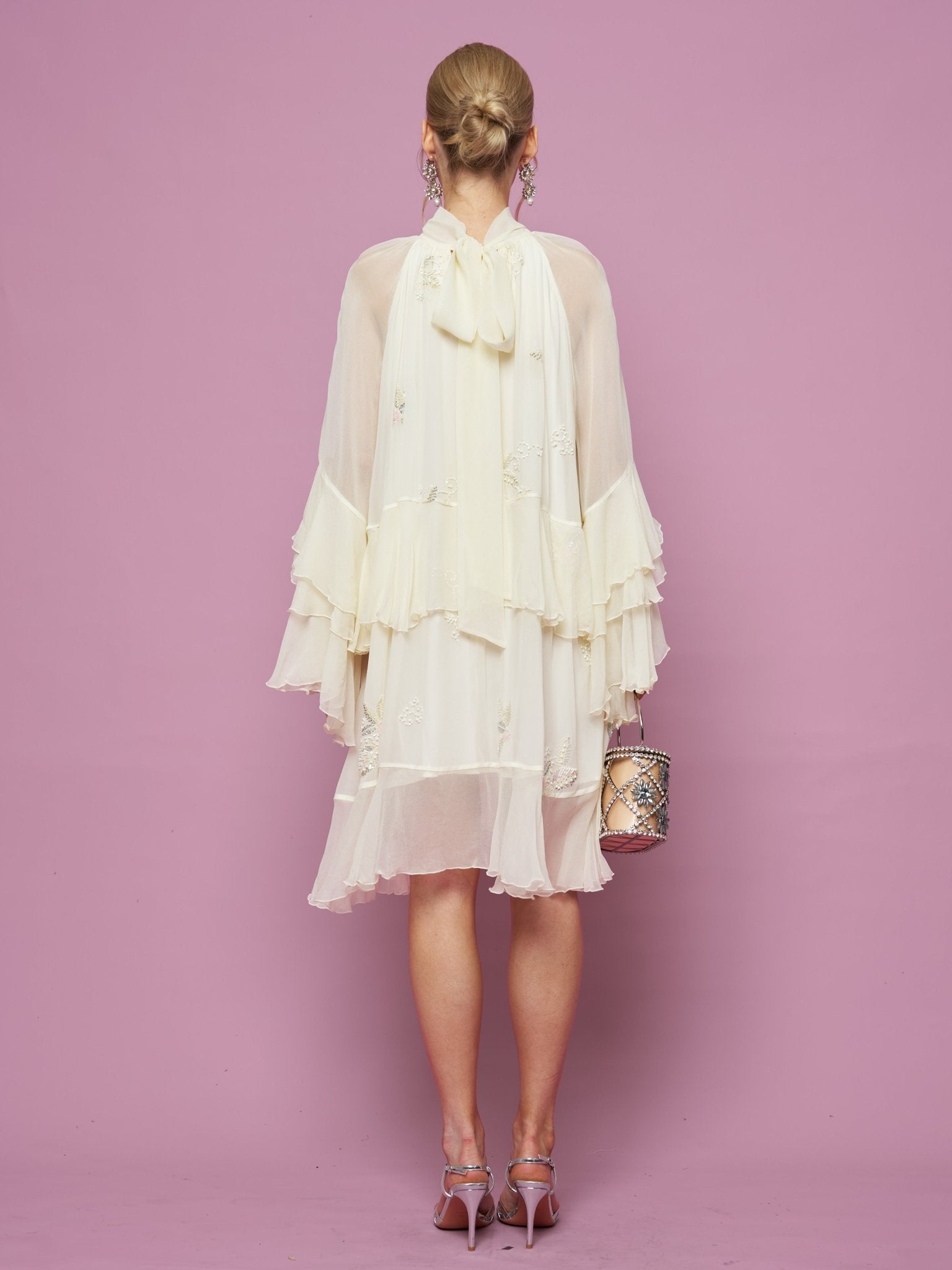 A woman faces a pink wall, wearing the Embroidered High-Neck Silk Dress by Social Queen—a cream-colored evening gown featuring cape detail and subtle sequin embroidery—paired with a small handbag and pink heels.