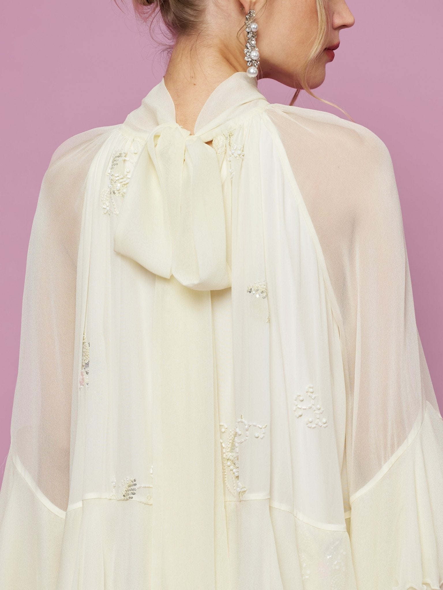 A woman shown from behind against a pink background wears the Social Queen Embroidered High-Neck Silk Dress in cream, featuring delicate sequin embroidery and a large back bow.