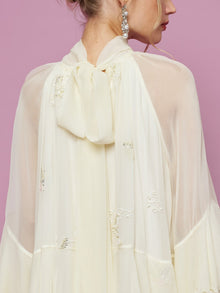 A woman shown from behind against a pink background wears the Social Queen Embroidered High-Neck Silk Dress in cream, featuring delicate sequin embroidery and a large back bow.