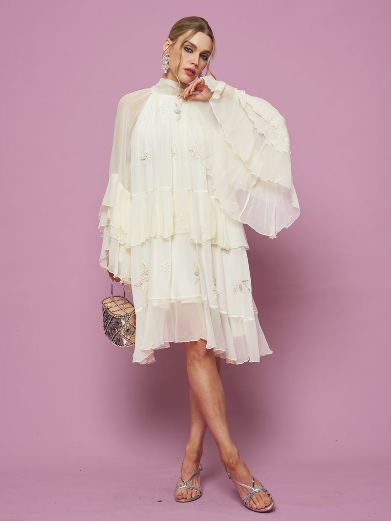 A person wears the Social Queen Embroidered High-Neck Silk Dress—white, knee-length with sheer sleeves and subtle sequin details—paired with sandals, statement earrings, and a small handbag against a pink background.
