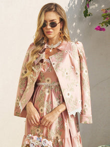 A woman wearing sunglasses and the Social Queen Embroidered Jacquard Lace-Trimmed Jacket draped over her shoulders, paired with a light pink floral dress, stands outdoors by a white wall with flowers in the background.