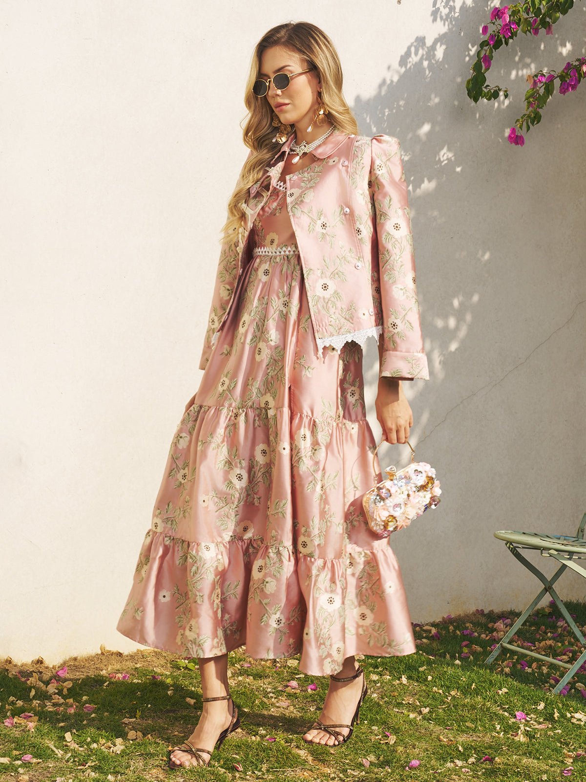 A woman stands outdoors near a wall and metal chair, wearing Social Queen's Embroidered Jacquard Lace-Trimmed Jacket with a matching dress and holding a small floral purse.