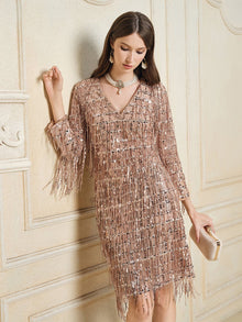 A woman wearing the Social Queen Embroidered Sequin Tassel Mini Dress in a neutral tone holds a clutch while standing against an ornate, light-colored wall.