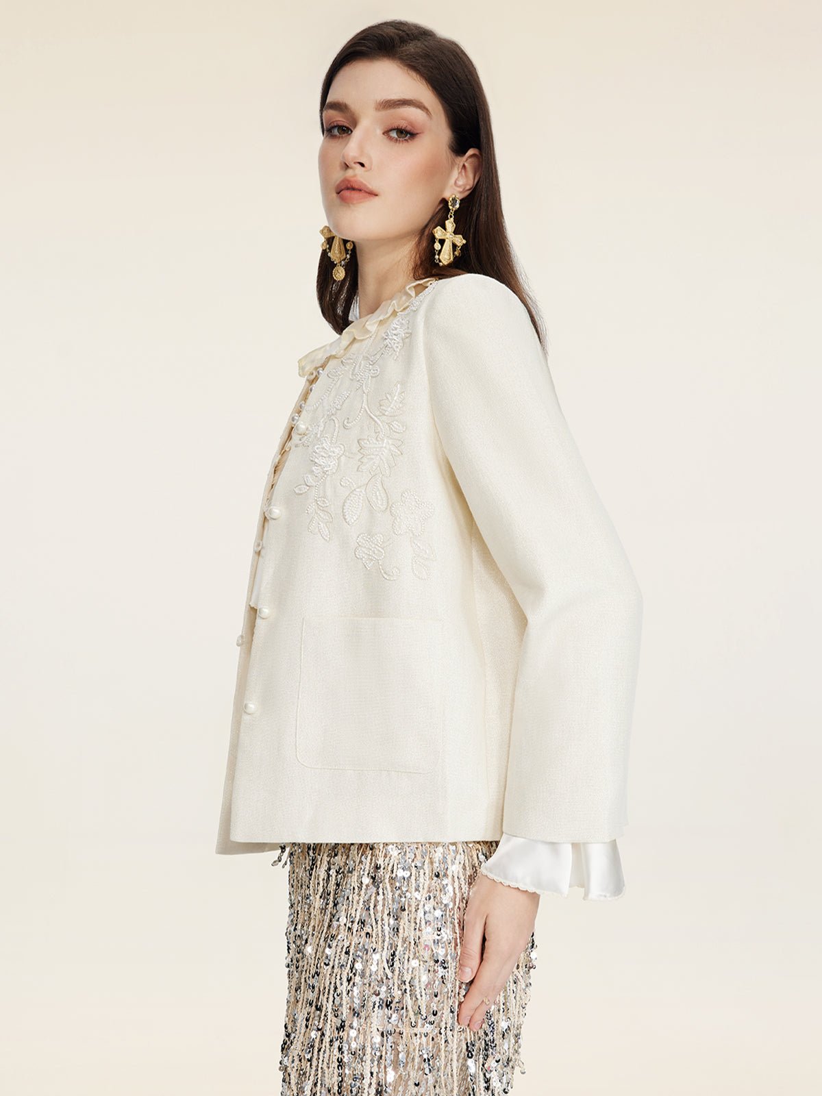 A woman in profile wears Social Queen's Embroidered Wool Jacket with Pearl Accents over a white ruffled blouse, styled with a metallic fringe skirt and gold statement earrings against a plain background.