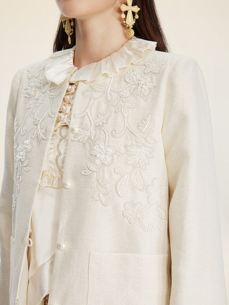 A woman wears the Social Queen Embroidered Wool Jacket with pearl accents, featuring a cream hue, hand-embroidered floral motifs, a ruffled collar, and gold floral earrings.