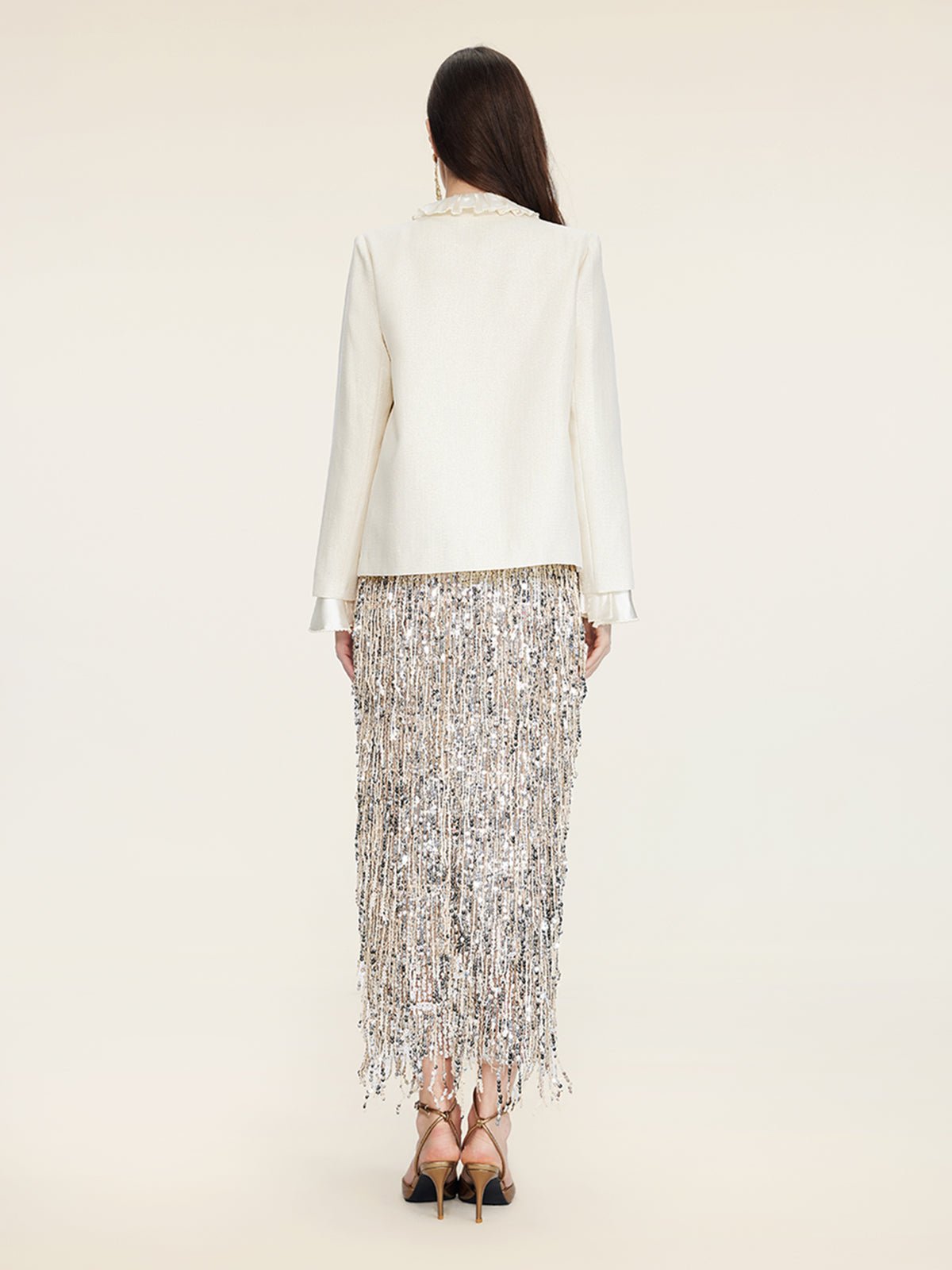 A woman faces away, wearing the Social Queen Embroidered Wool Jacket with pearl accents and a long silver sequin fringe skirt with high-heeled sandals, set against a plain background.