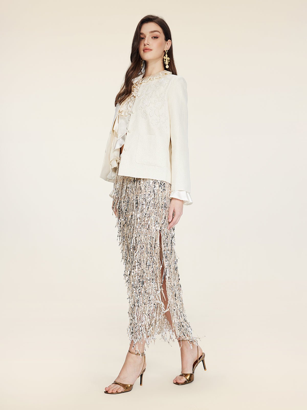 A woman in profile wears the Social Queen Embroidered Wool Jacket with Pearl Accents, a ruffled blouse, metallic fringe skirt, gold earrings, and high-heeled sandals against a neutral background.