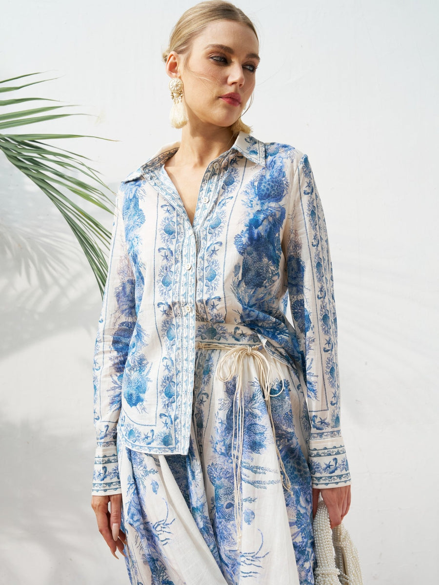 A woman wears the Social Queen Engineered Toile de Jouy Cotton Shirt in a blue and white floral pattern, paired with earrings and a rope belt, standing before a white wall with a plant in the background.