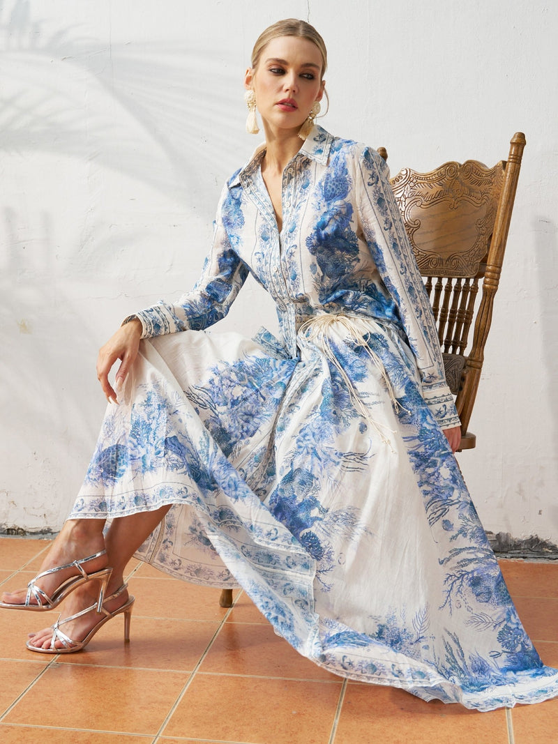 A woman sits on a wooden chair against a plain white wall wearing the Social Queen Engineered Toile de Jouy Cotton Shirt styled as a dress with silver high-heeled sandals.