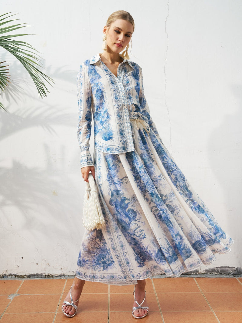 A woman stands against a white wall wearing the Social Queen Engineered Toile de Jouy Cotton Skirt, paired with a white fringed handbag, strappy sandals, and statement earrings.