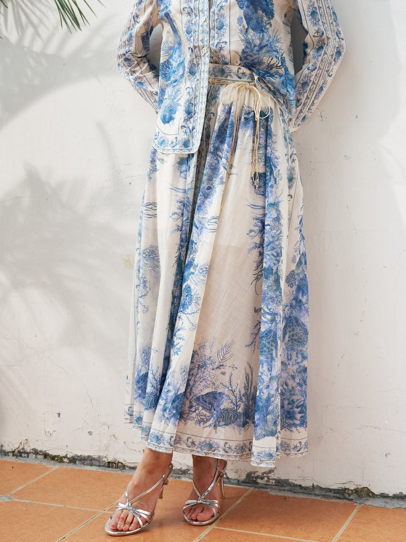 A person wears the Social Queen Engineered Toile de Jouy Cotton Skirt, featuring blue floral patterns and an adjustable drawstring waist, standing on a tiled floor against a white wall, paired with silver strappy sandals.