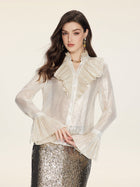 A woman wears the Social Queen Ethereal Metallic Ruffle Silk Blouse, featuring sheer silk with gold threads and a ruffled high collar, paired with a silver sequin skirt, against a plain light background.