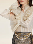 A woman models the Social Queen Ethereal Metallic Ruffle Silk Blouse, featuring gold threads, a high ruffled collar, and bell sleeves. She pairs it with a sequined skirt, layered pearls, and statement earrings. Her face is partially out of frame.