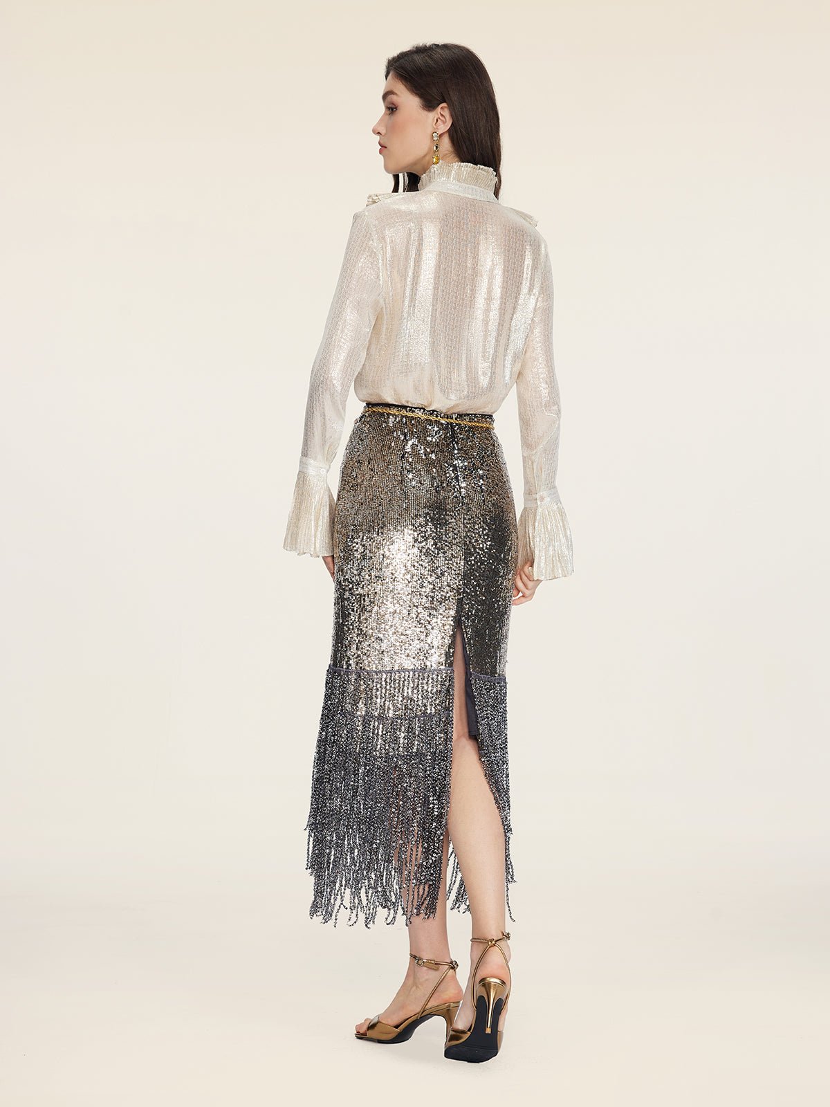 A woman stands facing away, wearing the Social Queen Ethereal Metallic Ruffle Silk Blouse, paired with a sequined fringe skirt and gold high-heeled sandals against a plain background.