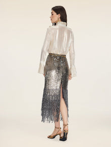 A woman stands facing away, wearing the Social Queen Ethereal Metallic Ruffle Silk Blouse, paired with a sequined fringe skirt and gold high-heeled sandals against a plain background.