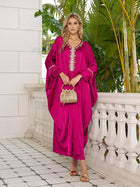 On a tiled balcony with greenery, a woman wears the Social Queen Feather-Trimmed Embellished Satin Caftan Dress, paired with gold sandals and a small beaded handbag.