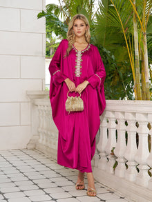 On a tiled balcony with greenery, a woman wears the Social Queen Feather-Trimmed Embellished Satin Caftan Dress, paired with gold sandals and a small beaded handbag.