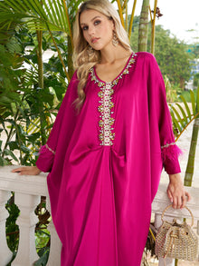 A woman poses outdoors near tropical plants and a white railing in the Social Queen Feather-Trimmed Embellished Satin Caftan Dress, paired with a beaded handbag.