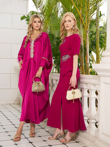 Two women in Social Queen Feather-Trimmed Embellished Satin Caftan Dresses stand by a white stone railing with green plants behind them. Each holds a small handbag and wears sandals.